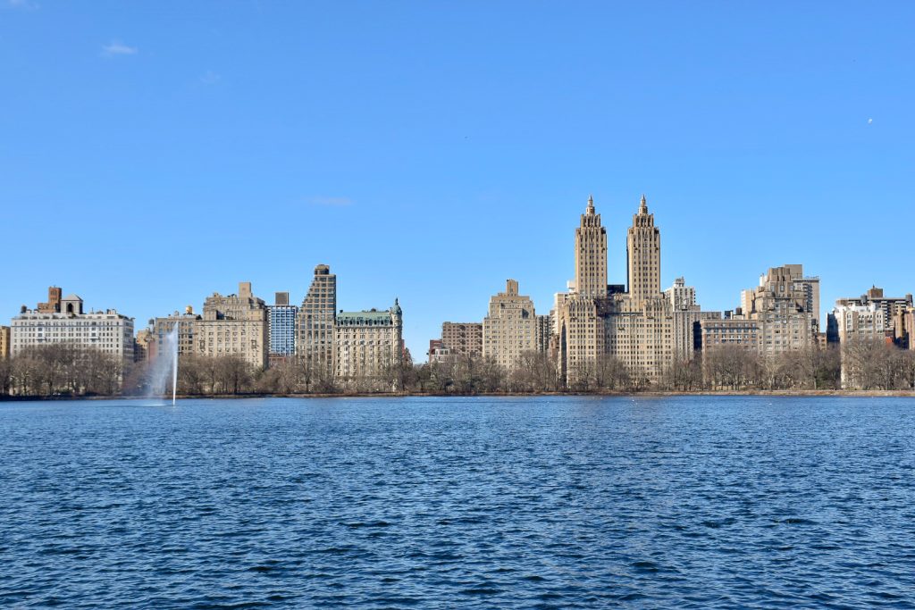 Line of smart buildings and towers across the middle of frame. Water in front and sky above. small water fountain to the left