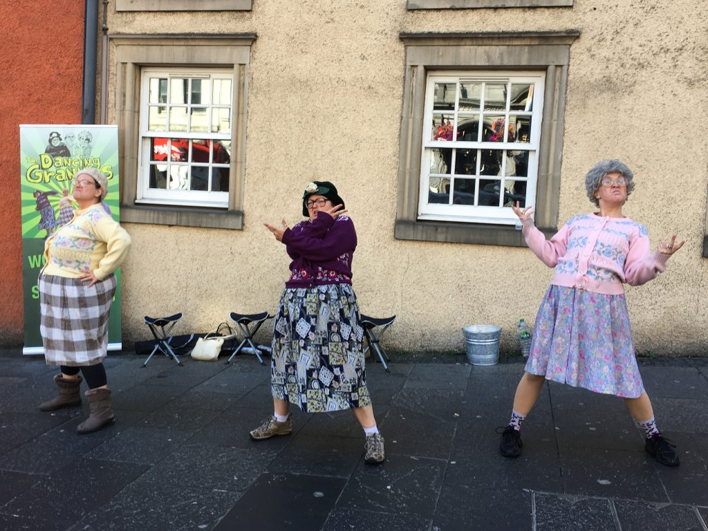Three men dressed as Granny's stand in a rap pose in front of a building with white framed windows. Looks comical
