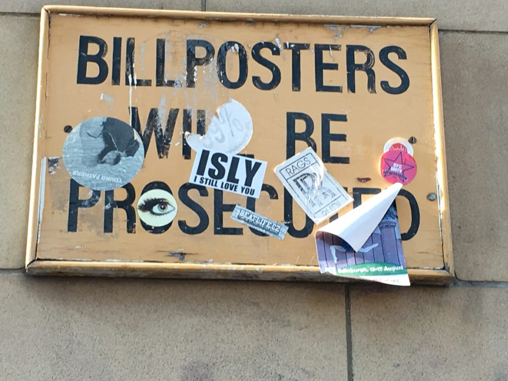 A beige notice board on the wall with black writing saying Bill Posters Will Be Prosecuted, but the sign is covered in stickers