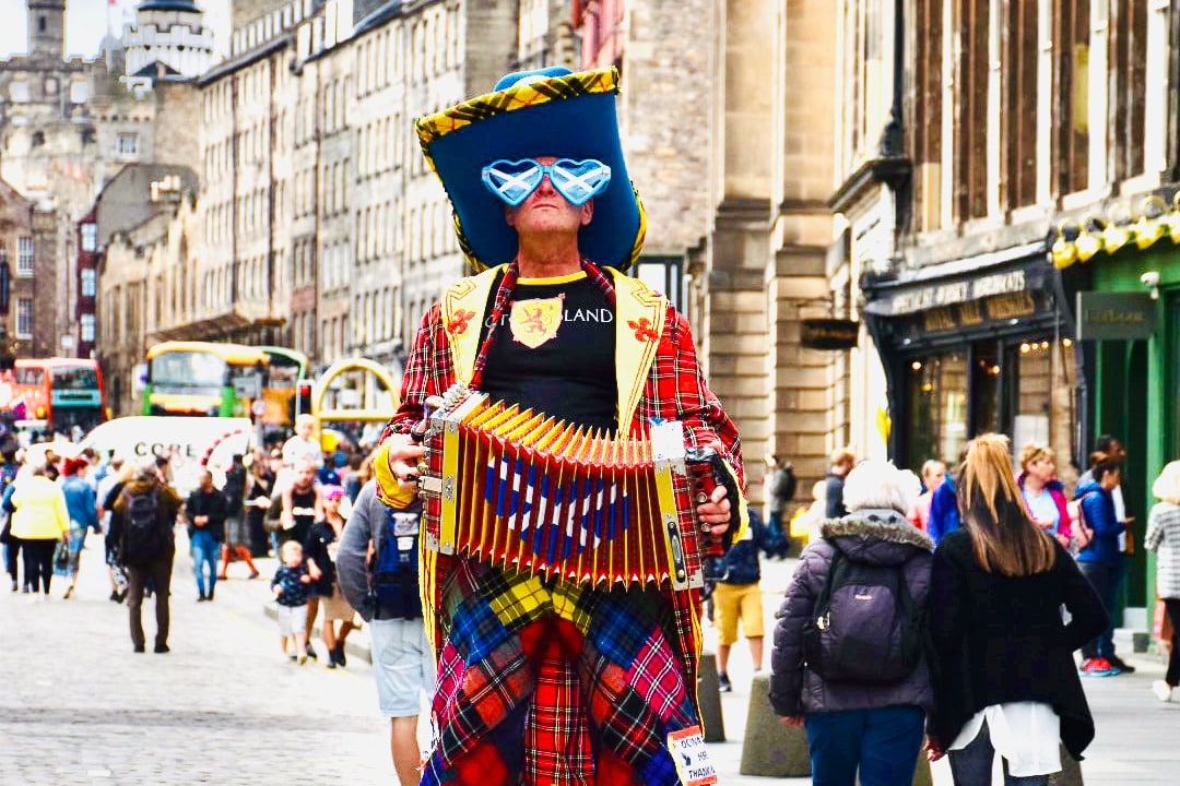 A man stands in the centre of the frame. He is wearing a big hat, glasses with the Scottish flag on the lenses, tartan trousers and a red coat. He is holding an acrodian open. Street scene behind the people wandering on road.