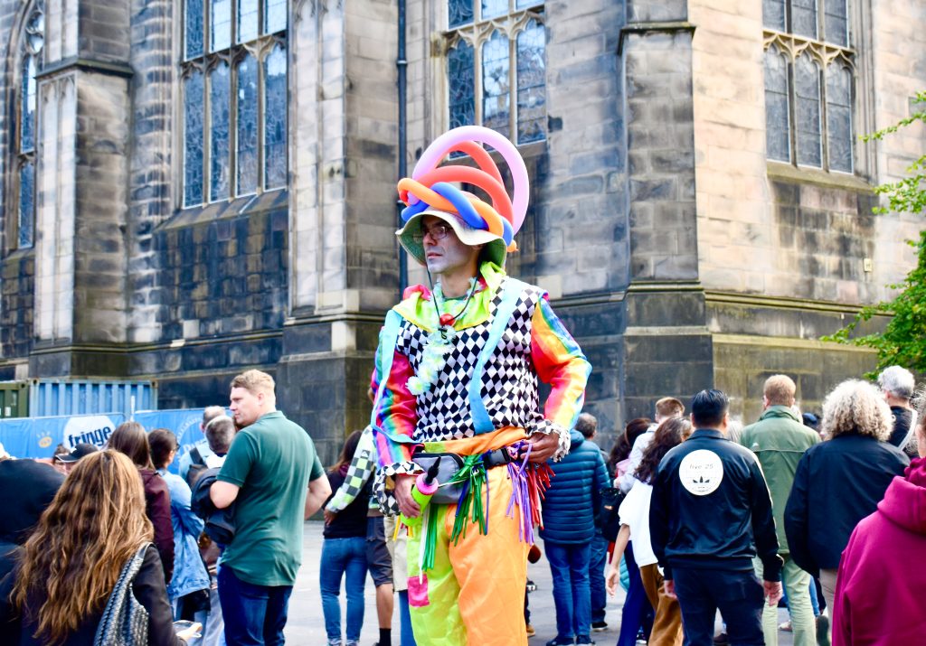 Man in yellow trousers, striped top and hat with long balloons on his head stands in the street with a brick building behind