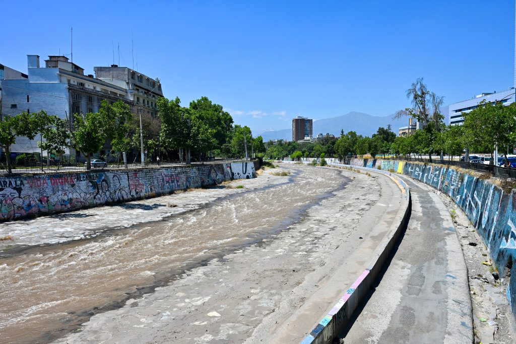 A half empty riverbed curves from the bottom left of the frame to the middle of the horizon. The river has concrete graffiti covered walls lined with trees and buildings behind. Faint mountains on the horizon. Blue sky above