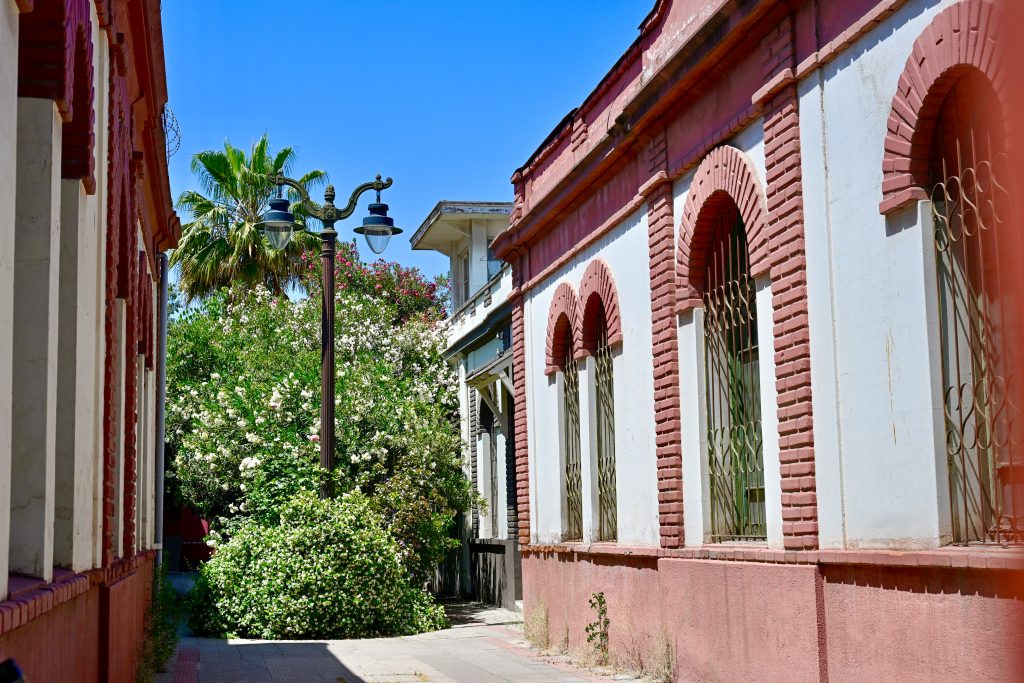 Looking between two pink and white buildings with green trees at the end of the alley. Black lamp post in the middle of the foliage. Pale blue sky above.
