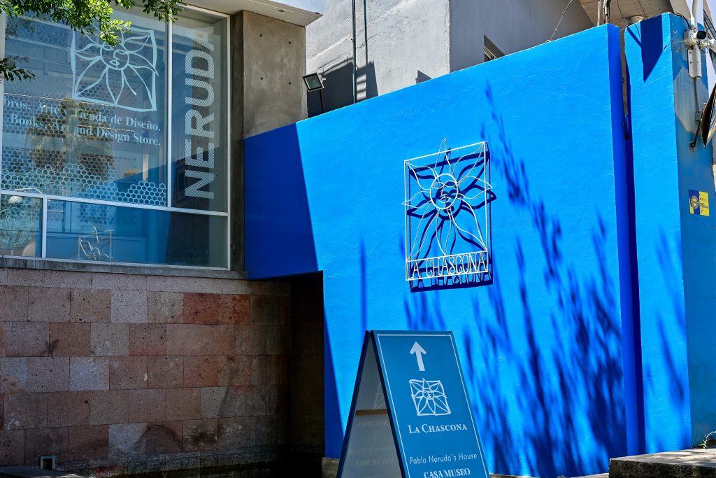 A blue painted wall fills most of the right hand side of the frame. It has a metal design saying La Chascona on ti and a sign advertising 'La Chascona, Pablo Neruda's House'. Glass window to the left advertising the Neruda museum