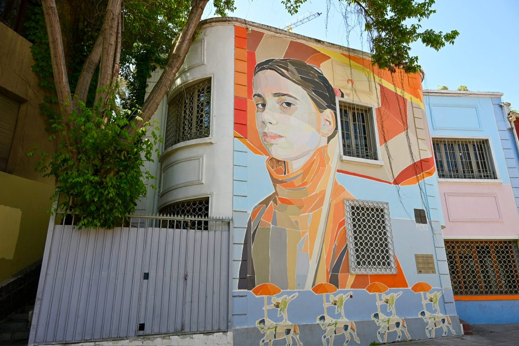 A two storey building sits in the middle of the frame. it has a large mural of a girl with brown hair, multicoloured coat and parasol, which fills the building. Tree to the left.