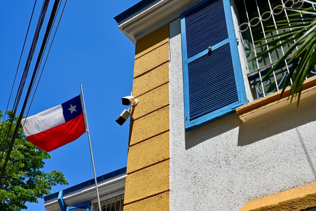Looking up at the side of a building. Chile flag to the left. Blue sky behind. The building is white with yellow trim and blue shutters on the window with iron bars.