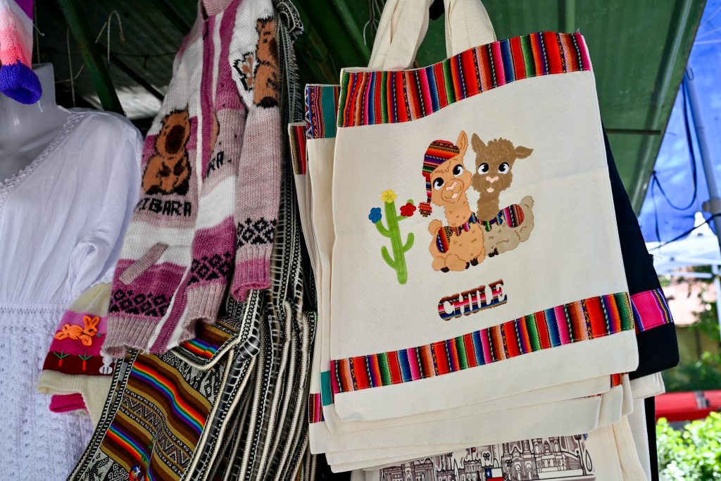 Items for sale hang from the roof of a market stall including, knitted jumper and bags. There is a cream bag featuring two llamas which says Chile on it