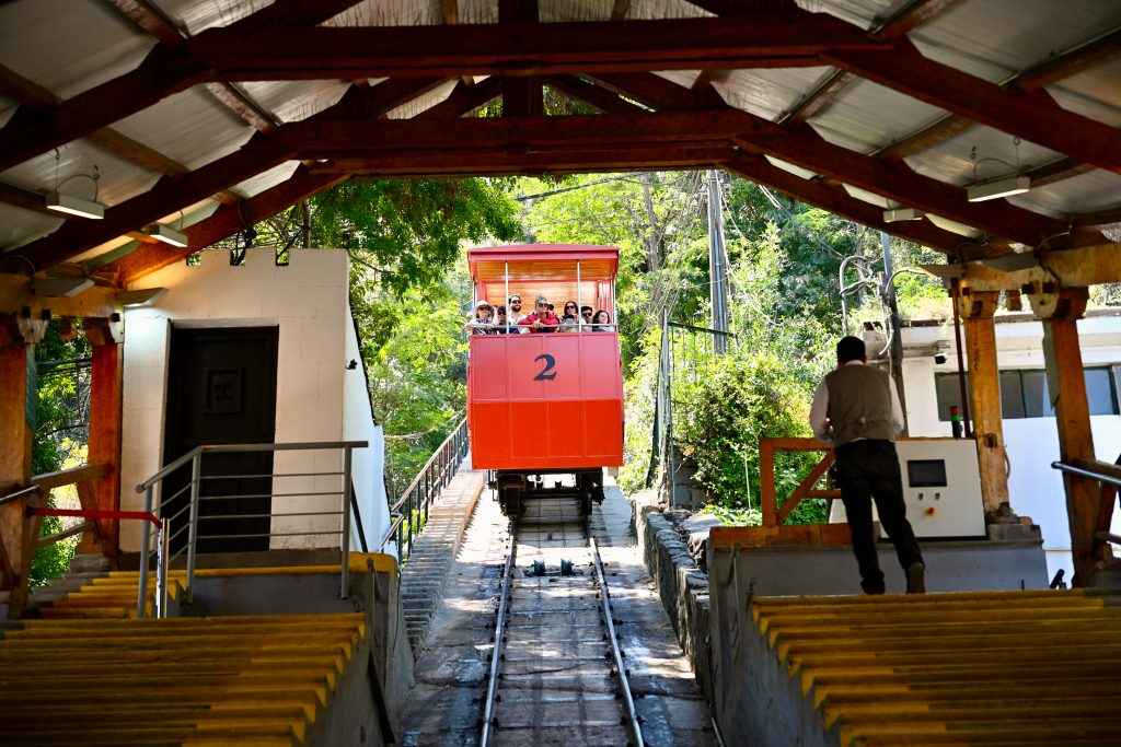 View looking up hill from inside a wooden garage. A red funicular car is approaching the camera on rails. There are passengers in the funicular car. A conductor on the ground to the right