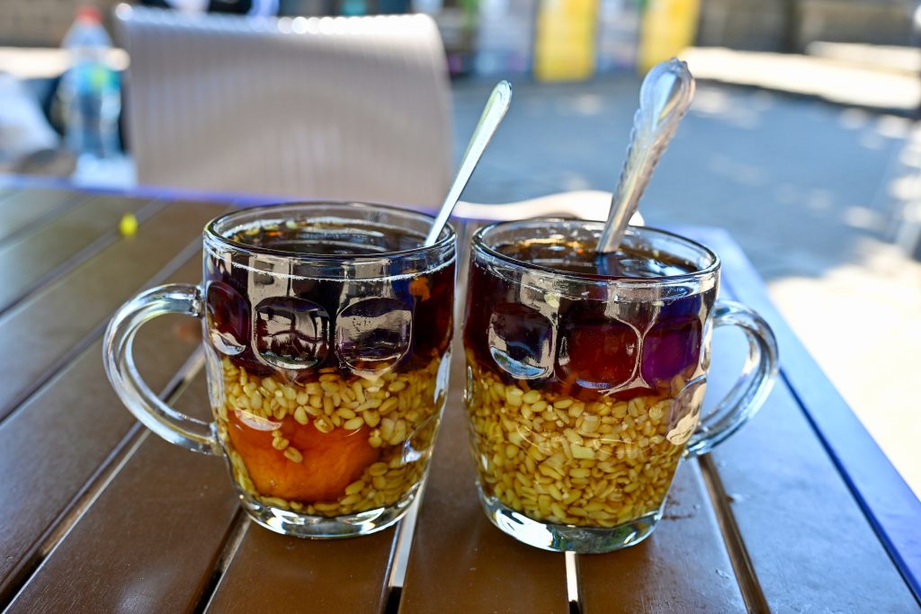 Two pint glasses sit on a brown table in an outdoors setting. They are filled with layers of what looks like fruit, oats and a dark liquid on top. A silver spoon in each glass.