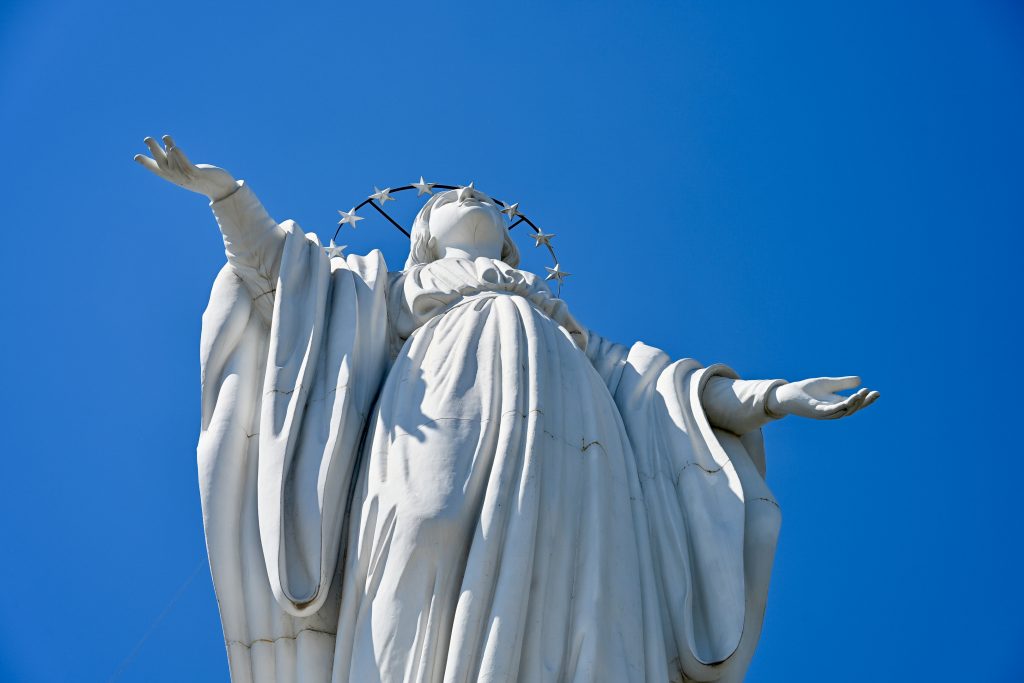 Looking up a clear blue sky. A white figurine statue fills the centre of the frame - looks like a lady in a flowing gown with halo of stars around her head. Arms outreached to the side
