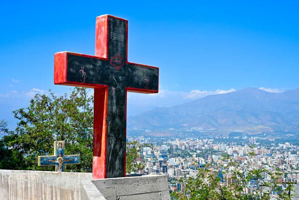 Sculpture of a black cross with red trim to the left of the centre of the frame. A second cross just visible to the left behind it. To the right is a city spread out across a valley with mountains behind. Pale blue sky above