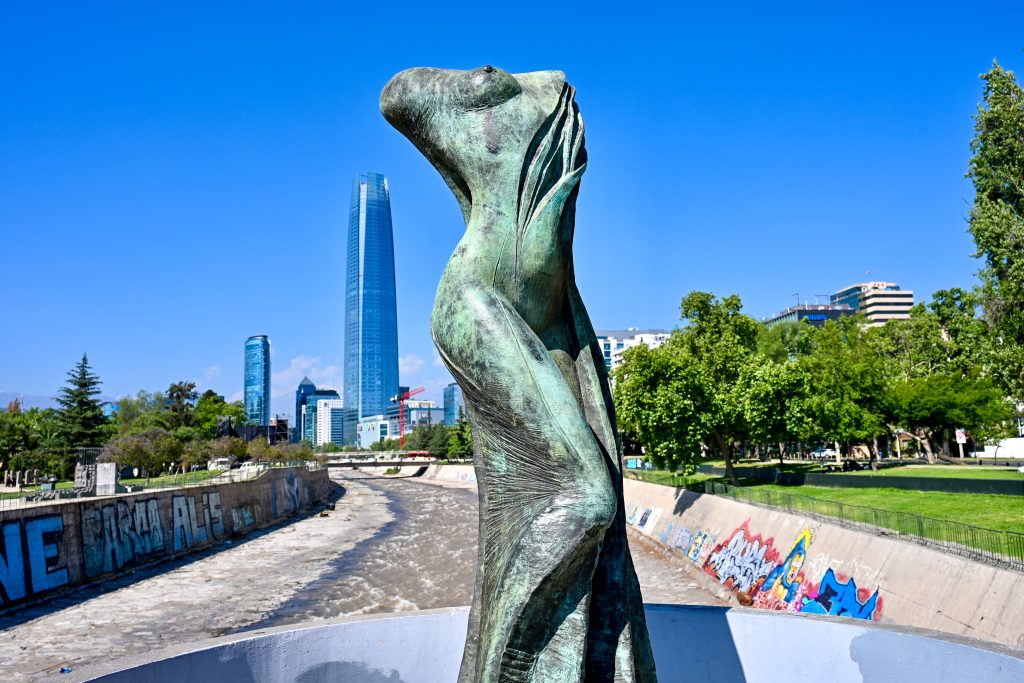 View along a half empty river bed towards modern blue skyscraper building on the horizon. Blue sky above. Green trees on the river banks. Graffiti on the concrete walls. A statue of a headless lady stands in the foreground.
