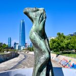 View along a half empty river bed towards modern blue skyscraper building on the horizon. Blue sky above. Green trees on the river banks. Graffiti on the concrete walls. A statue of a headless lady stands in the foreground.