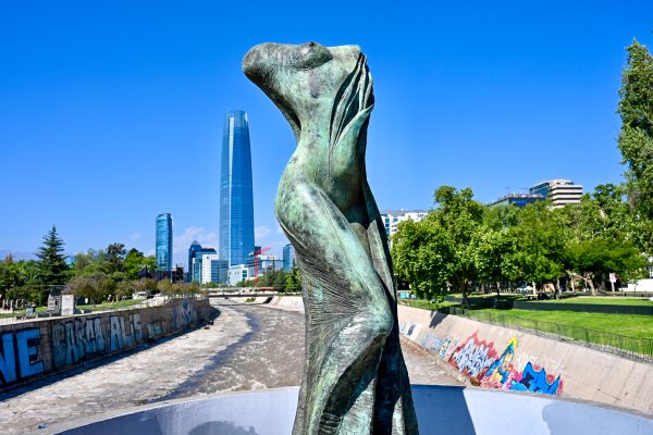 View along a half empty river bed towards modern blue skyscraper building on the horizon. Blue sky above. Green trees on the river banks. Graffiti on the concrete walls. A statue of a headless lady stands in the foreground.
