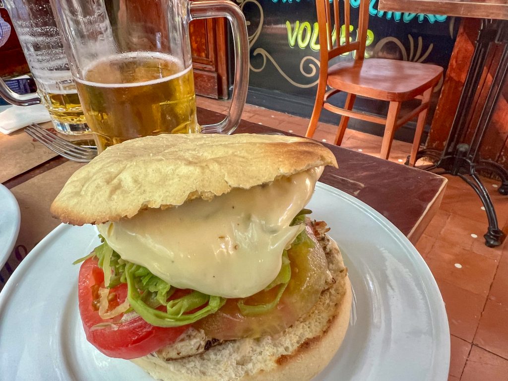View of a white plate sitting on a wooden table with pints of larger in the background. On the plate is a large sandwich with what appears to be chicken, tomato, green beans and cheese filling).