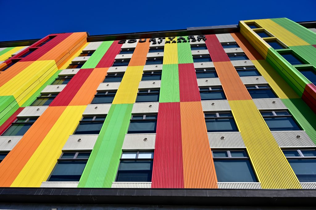 Looking upwards to the top of a hotel building with square windows. Flat roof. Dark blue sky above. The hotel is decorated with coloured stripes of yellow, red, orange and green blocks between the windows. It says Courtyard Marriott' at the top