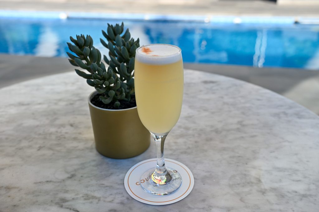 A tall glass of yellow liquid with a white frothy top sits on a white coaster on a circular marble table next to a green plant. Swimming pool behind