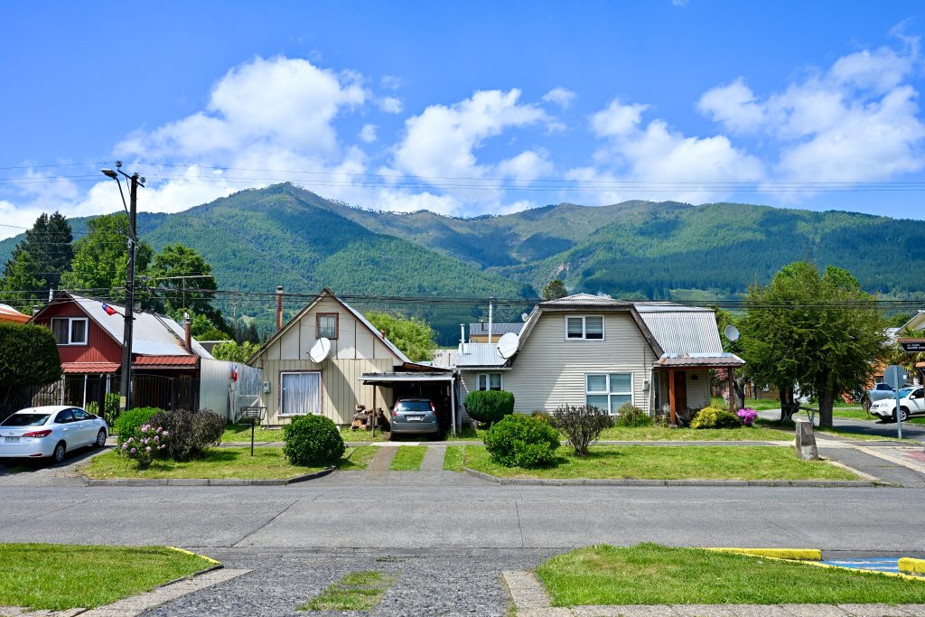 A row of 3 wooden houses sits on the horizon. They have cars in the drive with grass verges, shurbs and trees in front. A road in the foreground. Tree covered hills behind with blue sky above