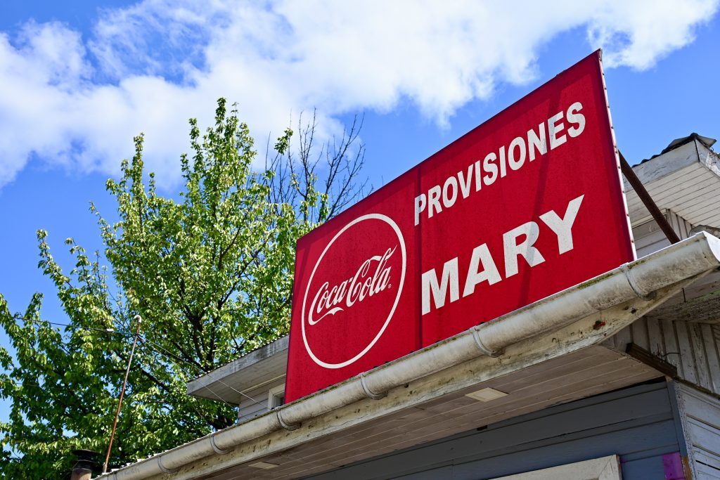 Looking up at a red sign on a grey wooden building to the righthand side of the frame. There is a tree to the right and blue sky with white cloud above. The red sign says 'Provisiones Mary' in white and also has the Coca-Cola logo to the left