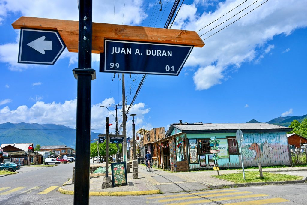 View of a signpost at a cross roads on a street with hills in the background. The signpost says 'Juan A Duran' on it and points to the left. There are yellow stripes on the road. A row of single storey wooden buildings on the other side of the road