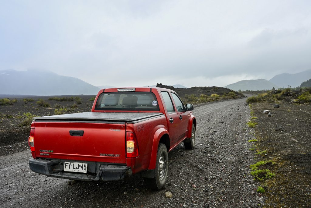 A red pick up truck stands in the bottom left of the frame on a gravel road surrounded by black volcanic rocs with small green shrubs. Cloudy sky above