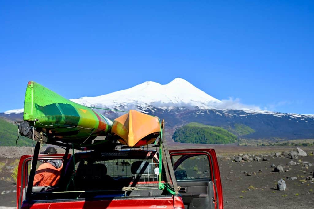 A red pick up truck is parked in the bottom left corner of the frame. Its doors are open and it has a green and orange kayak attached to its roof. Black volcanic soil behind and a snow capped volcano on the horizon with a double cone. Blue sky above