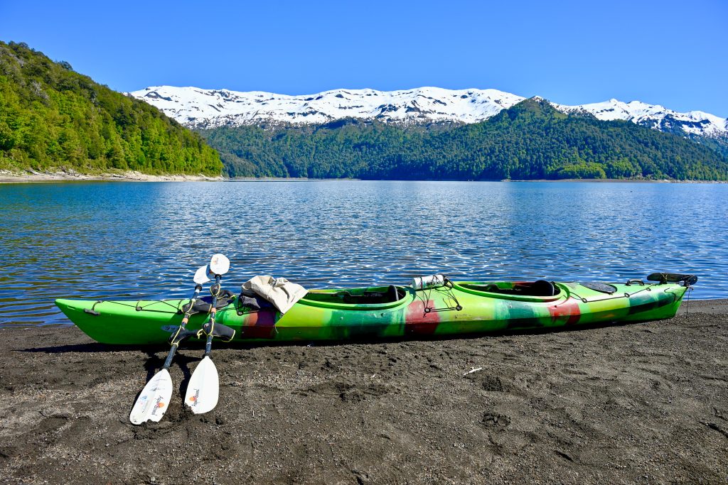 Top third of the frame is a row of snow-capped mountains in front of tree covered hills. The middle third is a flat blue lake. Bottom third is black volcanic soil with a green double kayak parked on the water's edge. Two white oars sit expectantly on the side of the kayak