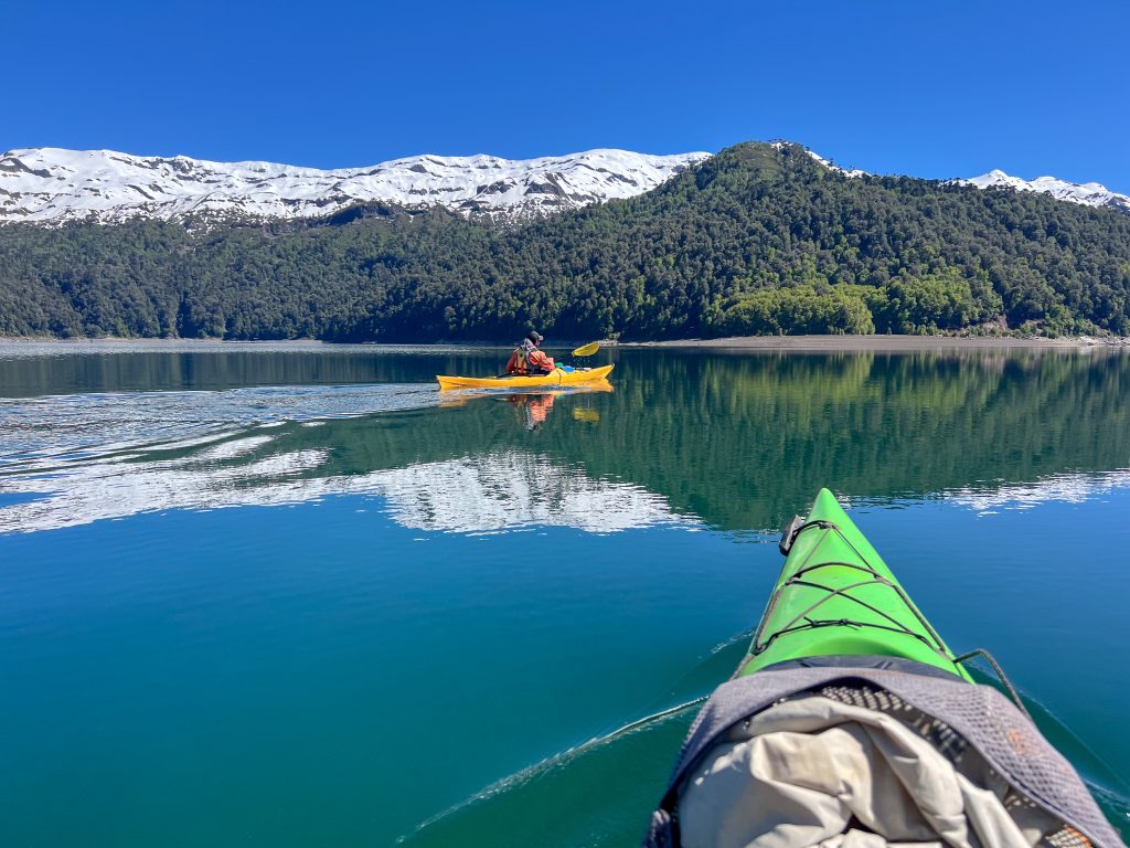 A beautifully flat lake across the two-thirds of the frame. A row of tree covered hills above the lake with snow-covered mountains behind and deep blue sky above. The mountains are reflected in the water of the lake which acts like a mirror. The nose of a green kayak enters the frame from the bottom right and a yellow kayak makes its way across the lake from left to right in the middle of the frame