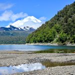 Landscape view of a gravel sandbank in the foreground leading to a flat blue lake. Trees and a large snow-capped volcano behind. Blue sky above. Tree covered hill to the right