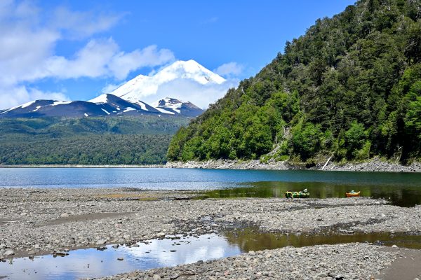 Landscape view of a gravel sandbank in the foreground leading to a flat blue lake. Trees and a large snow-capped volcano behind. Blue sky above. Tree covered hill to the right