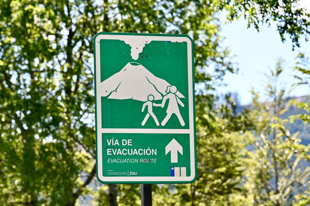 Trees in background with pale blue sky behind. Upright rectangular green sign in centre of frame with white diagram of volcano and two graphic people walking to the right. It says "Via de Evacuation / Evacuation Route" a the bottom