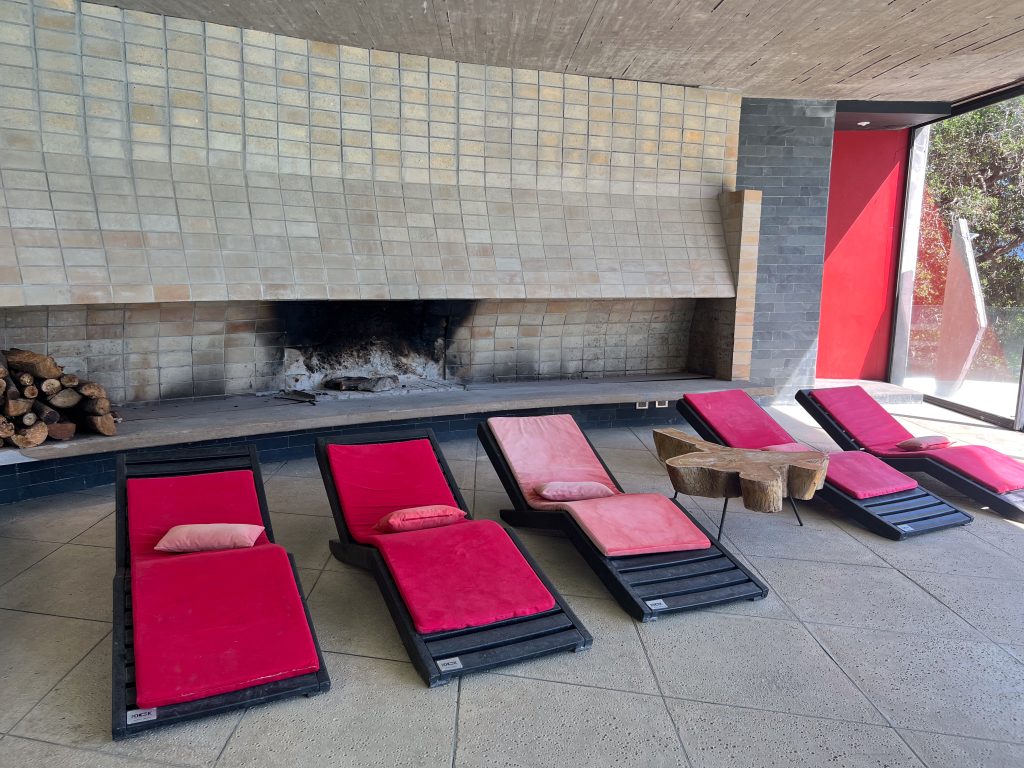 A row of black sun loungers crosses the front of the shot. Each sunlougner has a red mattress and a red cushion on it. Behind is a tiled wall above a real fire with pile of logs to the left. Window to the right hand side