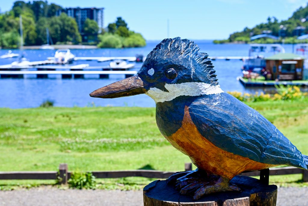 A carved wooden kingfisher sits I the bottom right of the frame. It's painted blue white and orange. Behind it is a lake with small wooden piers, trees and a hotel- out of focus. Pale sky above