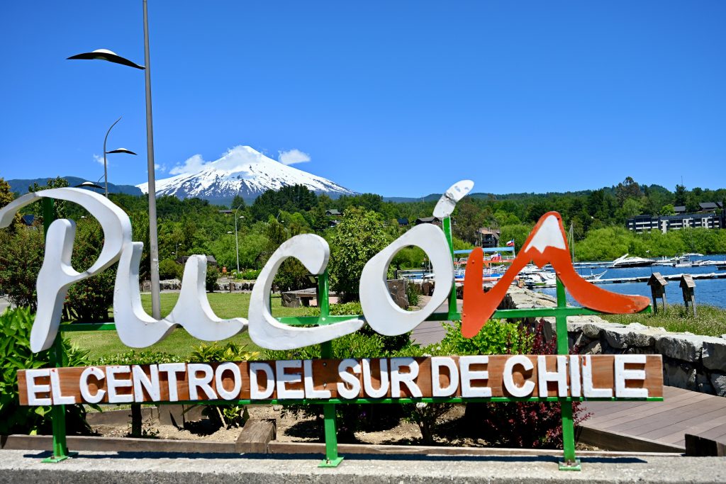 A white sign runs across the bottom of the frame which says 'Pucon, El Centro Del Sur de Chile'. The N on Pucon is shaped like a volcano in red. Behind is a park with signs of a lake to the right. In the distance behind a row of trees is the cone of a snow-capped volcano. Blue sky above