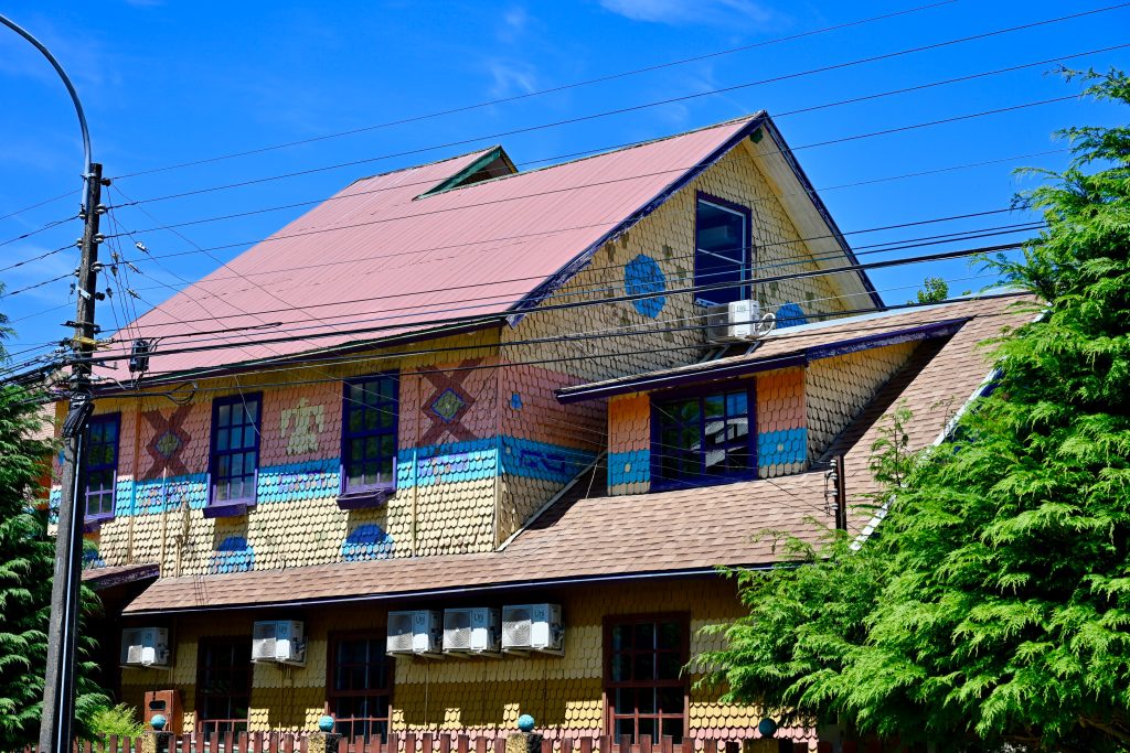 A wooden building fills the frame. It has a red roof and multicoloured wooden panels all over the building - yellow, orange, blue and some geometric patterns. Blue sky above. Tree to the right. Lamppost to the left