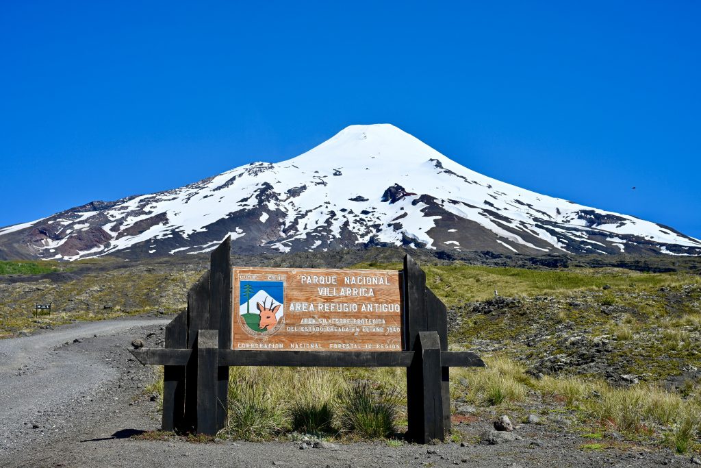 View across a patch of land covered with stones and grass, towards a large snow capped volcano on the centre of the horizon. here is a wooden sign in the centre foreground saying 'Parque Nacional Villarricca. Area Reugio Arntigo'. It has a shield with picture of a deer on it