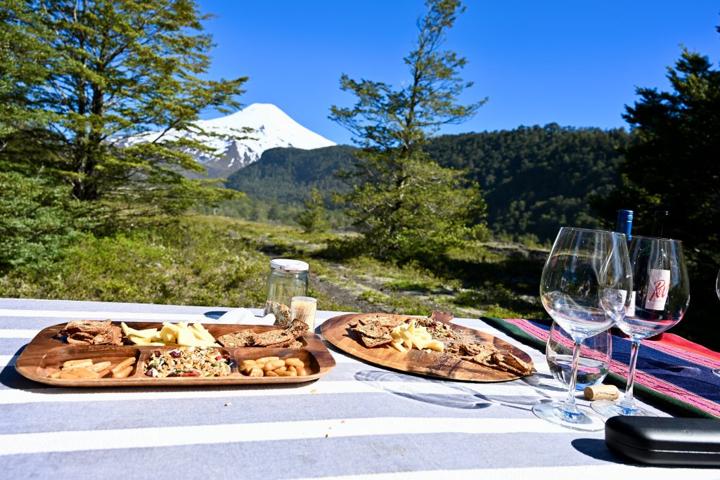 View over a table covered in blue and white table cloth towards green hill, trees and a snow-capped volcano on the horizon. Blue sky above. There are 2 plates of snacks on the table and 2 white glasses to the right hand side
