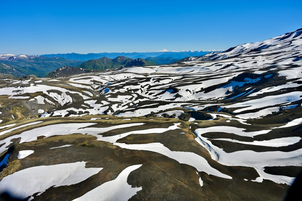 View across a mountain side of land with dark soil covered with stripes of snow. Hills and mountains on the horizon. Blue sky above