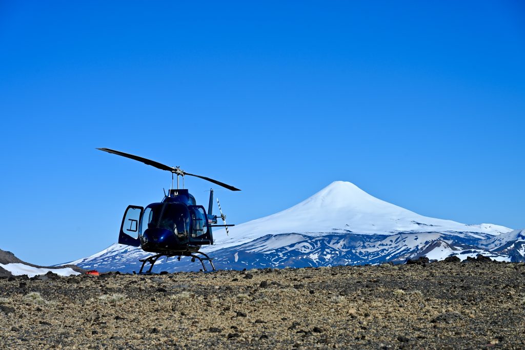 A strip of rocky land sits across the bottom of the frame. A black helicopter sits on the ground the left of centre. A large snow called volcano can be seen in the background. Clear blue sky above