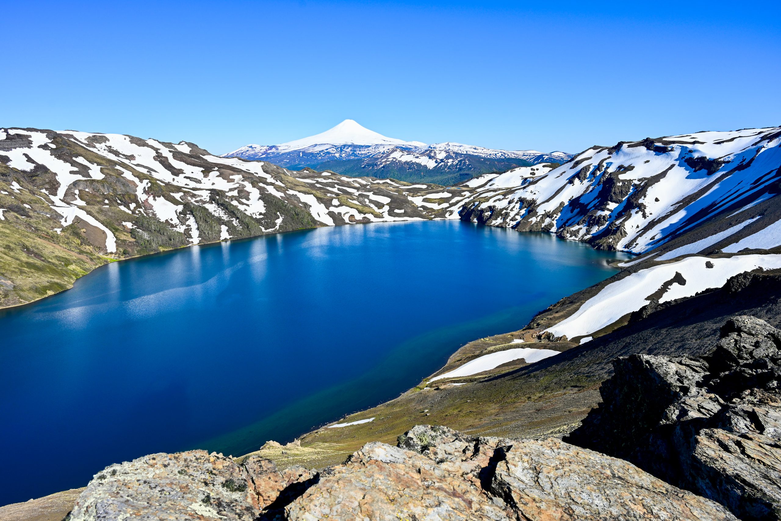 View from the top of a rocky hill looking over an oval blue lake encircled by snow covered rocks. The cone of a snow-capped volcano sits on the horizon. Pale blue sky above