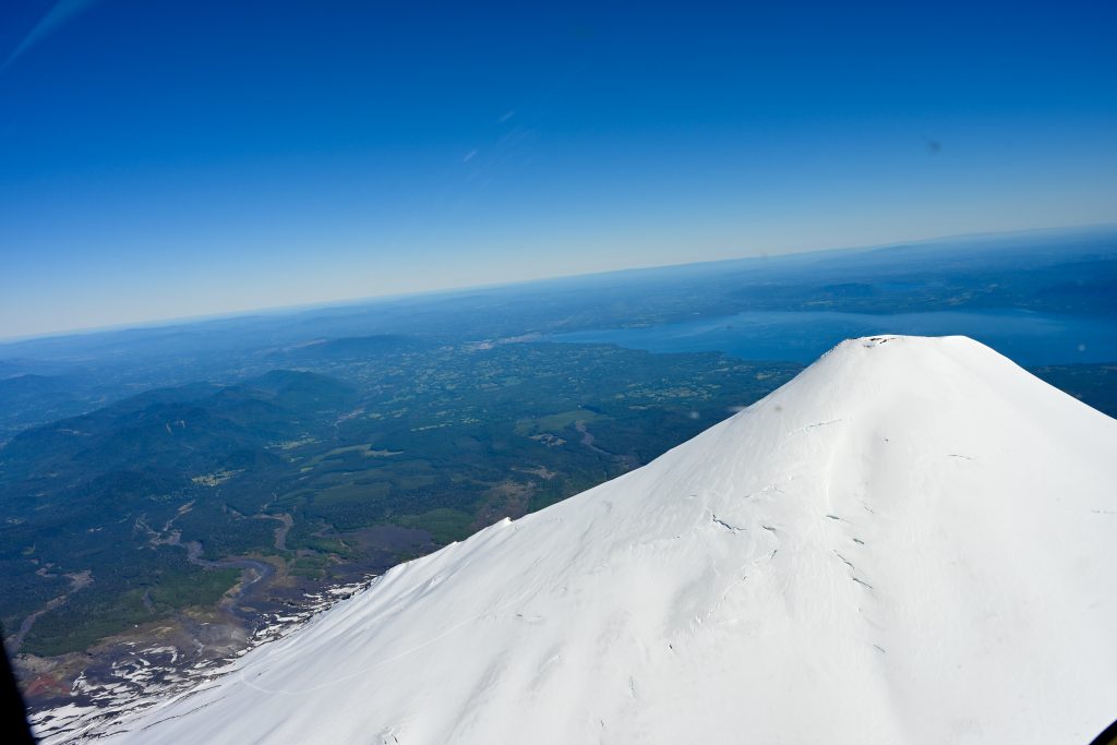 A volcano cone rises from the bottom right of the the frame. It is covered in snow. A valley filled with trees and a lake lie behind the volcano. Blue sky above