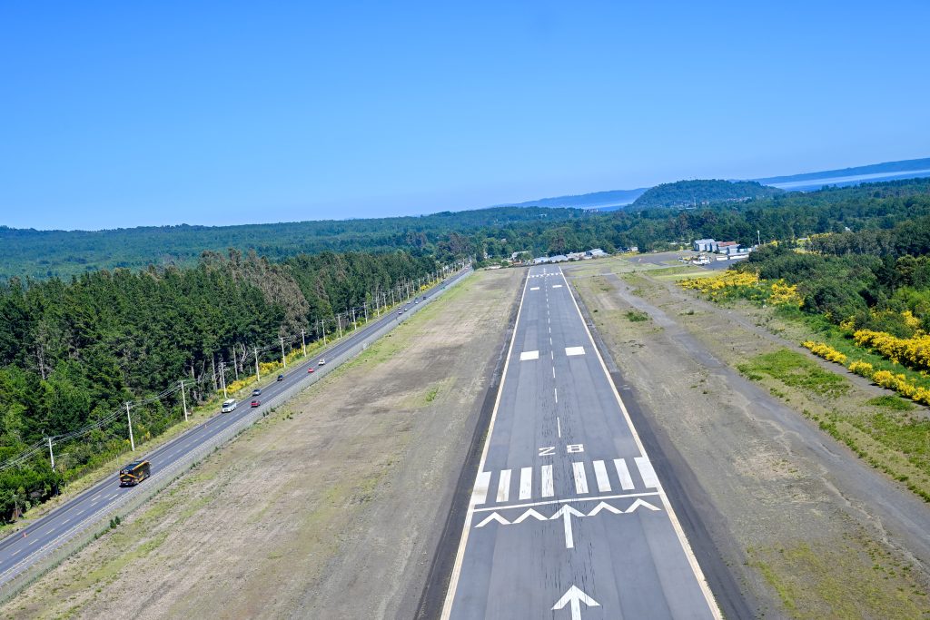 View from above of a short airport runway with trees around and a lake on the horizon. Blue sky above