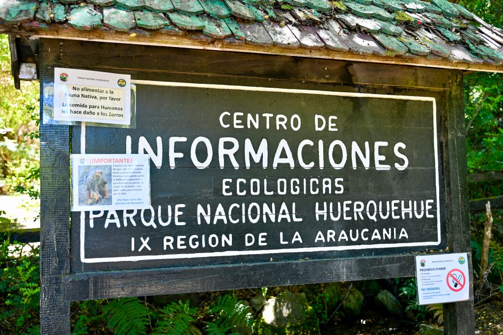 A wooden sign post fills the frame. It is black with a green roof and says 'Centro de Informaciones, Ecological, Parque Nacional Huerquehue' on it. There are three which signs stuck on top of the signpost