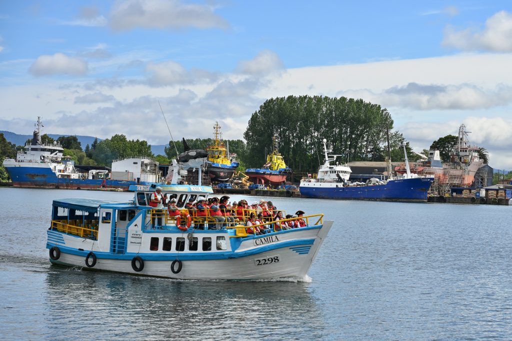 A white boat with turquoise trim enters the frame from the bottom left hand side. It is filled with sightseers sitting on the outdoor section at the front of the boat. they are wearing pirate hats. On the far side of the river are a number of boats moored in the river and also in the dry dock