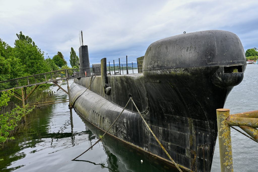Looking down the length of an old black submarine. There is a gangway to allow people on to the sub from the left hand side. Trees to the left. Grey clouds above. Grey water below