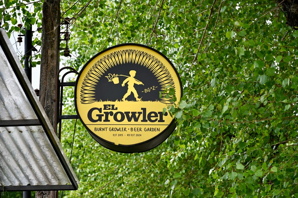 Looking up at a circular yellow sign featuring the diagram of a boy carrying a bucked on a stick over his shoulder. It is surrounded by flower petals almost like a sunflower. Underneath it says 'El Growler - Burnt Growler, Beer Garden'. Trees behind. Corregated roof to the left