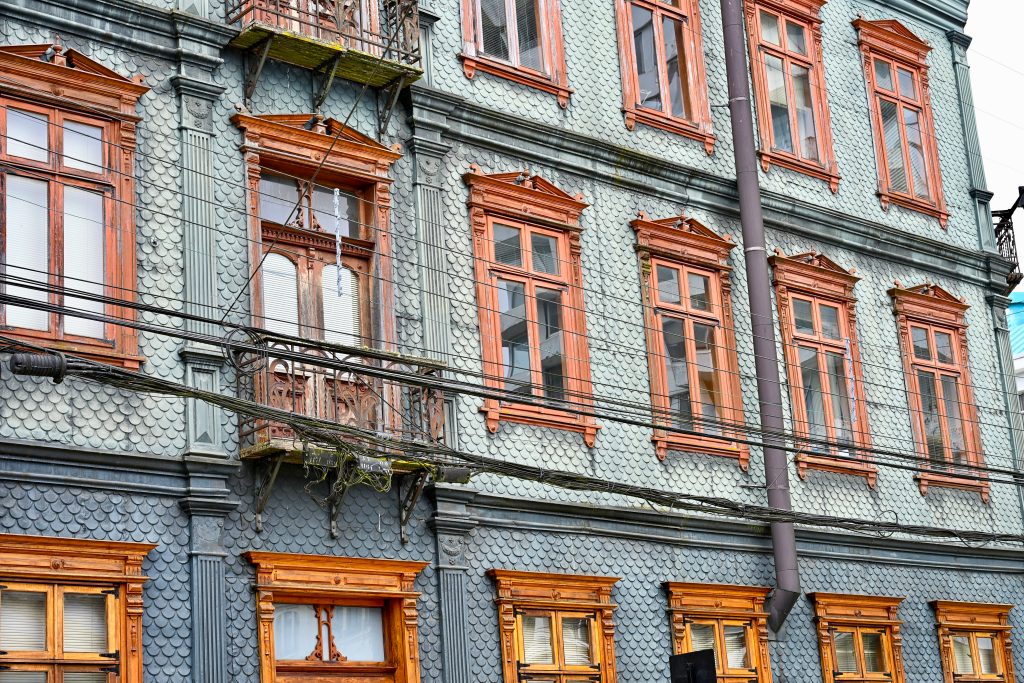 Looking up at the facade of a large building covered in turquoise tiles shaped like the scales of a fish. There are 3 storeys in shot each with a line of wooden window frames. One of the windows has an iron balcony to the middle left of the frame. There are wires running in front of the building