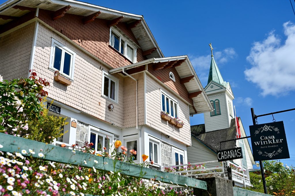 Looking up at a wooden building with double triangular roof and pale pink tiles on the building. Flowers in front bottom left. White church spire to the right. Signs saying Cabanas, Casa Winkler to the right
