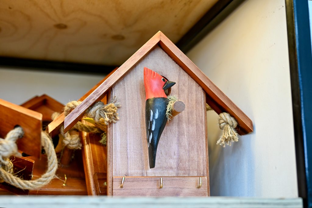 A wooden house with a woodpecker on the front sits on a shelf. The woodpecker has a black body and red head. It has hooks on the bottom as if for holding keys