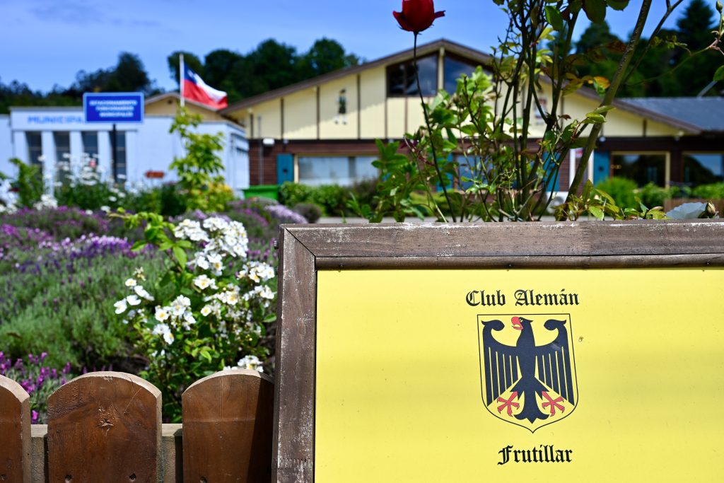 View over a wooden fence with a yellow sign saying Club Aleman, Frutillar on it with a German motif. Behind is a garden with white and purple flowers, a chalet building and Chile flag